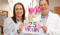 A woman and a man in a white doctor's coat smiling and  holding up a sign that says 25 years with pink and yellow balloons.