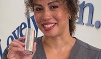 A woman with a medium skin tone and curly brown and blonde hair smiles while holding Dermalogica Smart Density Undereye Serum.
