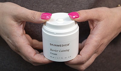 A person with pink fingernails holds a jar of SkinMedica Barrier Calming Cream while pressing down on the airless pump jar to dispense product.