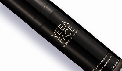 A black bottle of Veea Face Brightening Serum lays on a white background. The label on the bottle is in gold.