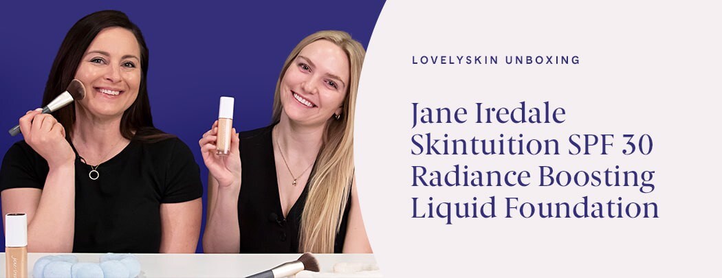 A woman with dark brown hair and light skin holds a makeup brush to her cheek while a blonde woman with fair skin holds a bottle of foundation.