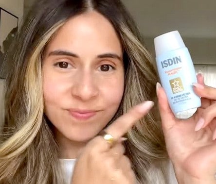 A woman with a medium skin tone and dark brown hair smiles as she points to a blue bottle of ISDIN Fusion Water Magic Broad Spectrum SPF 40 sunscreen.