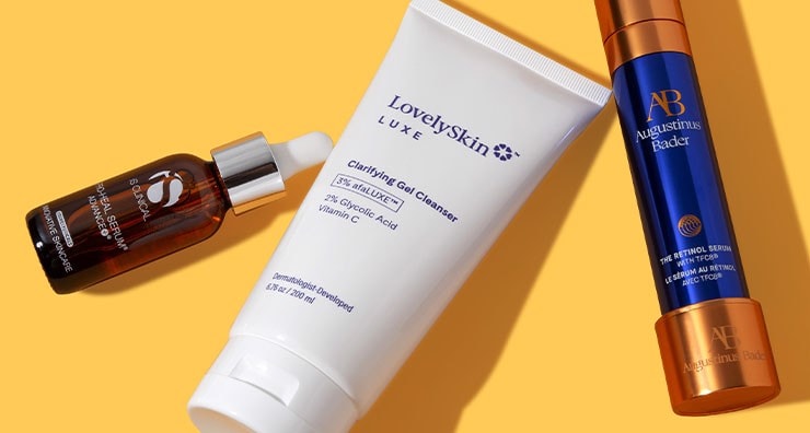 A white tube of LovelySkin LUXE cleanser and bottles of face serum from iS CLINICAL and Augustinus. Bader on a light orange background.