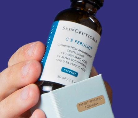 A person with an olive skin tone holds a bottle of SkinCeuticals C E Ferulic, almost as if they're pulling it out of the box.