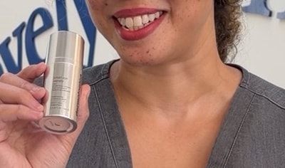 A woman with a medium skin tone and curly brown and blonde hair smiles while holding Dermalogica Smart Density Undereye Serum.
