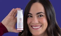 A woman with medium skin tone and dark hair holds a white bottle of EltaMD UV Clear Blemish-Prone & Oil Balancing SPF 50 near her face while smiling.
