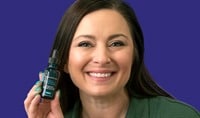 A woman with light skin and dark brown hair smiles at the camera while holding a bottle of SkinCeuticals A.G.E. Interrupter Ultra Serum.