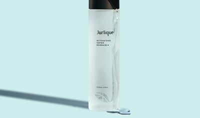A clear bottle of Jurlique Activating Water Essence+ with liquid running over the bottle and black cap on a soft blue background.