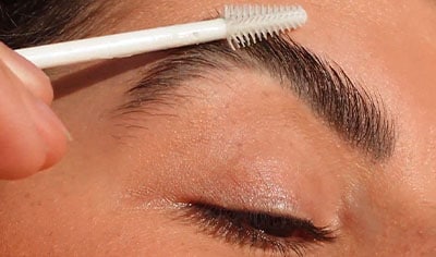 A person with medium, olive-toned skin holds a Ere Perez Aloe Gel Lash & Brow Mascara brush to their eyebrow.