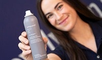 a woman with a medium skin tone and dark brown hair holds a bottle of Living Proof Dry Shampoo to the camera while smiling.