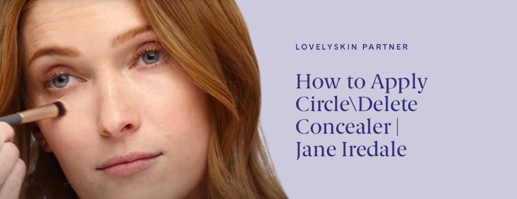A woman with fair skin and red hair uses a makeup brush to apply Jane Iredale Circle\Delete Concealer beneath her eyes.