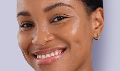 A woman with a deep skin tone smiles at the camera. Her skin looks dewy and radiant.