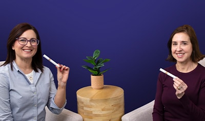 Two women, one in a dark purple shirt and one in a light blue shirt, hold Jane Iredale Skintuition Brightening Under Eye Concealer while smiling.