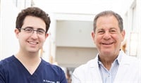 Drs. Joel and Daniel Schlessinger stand next to one another smiling. Dr. Daniel is wearing navy blue scrubs. Dr. Joel is wearing a white coat.