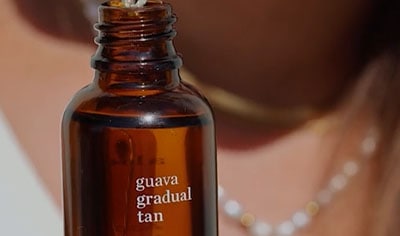 A person with a light to medium skin tone holds a bottle of Ere Perez Guava Gradual Face Tan with the dropper hovering over the open bottle.