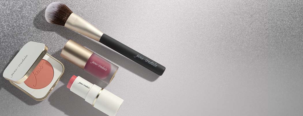 A powder blush in a compact, a cream blush in a tube, and a liquid blush in a tube lay next to a blush brush.