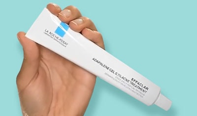 Hand holding white tube of La Roche-Posay Effaclar Adapalene Gel 0.1% Acne Treatment on light blue background