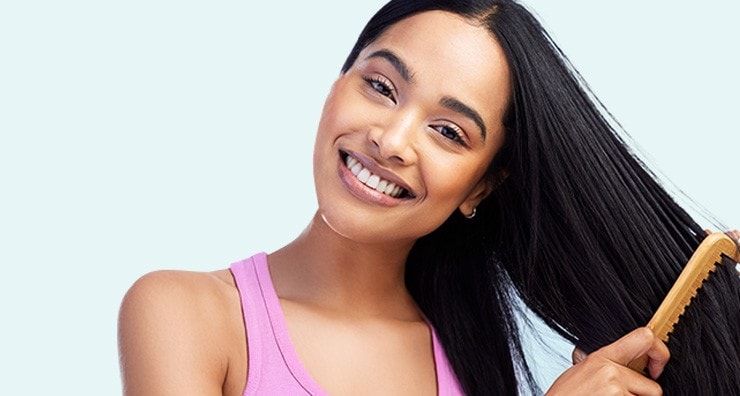 A female-presenting model with tan skin and straight black hair smiling and combing her hair with a wooden comb.