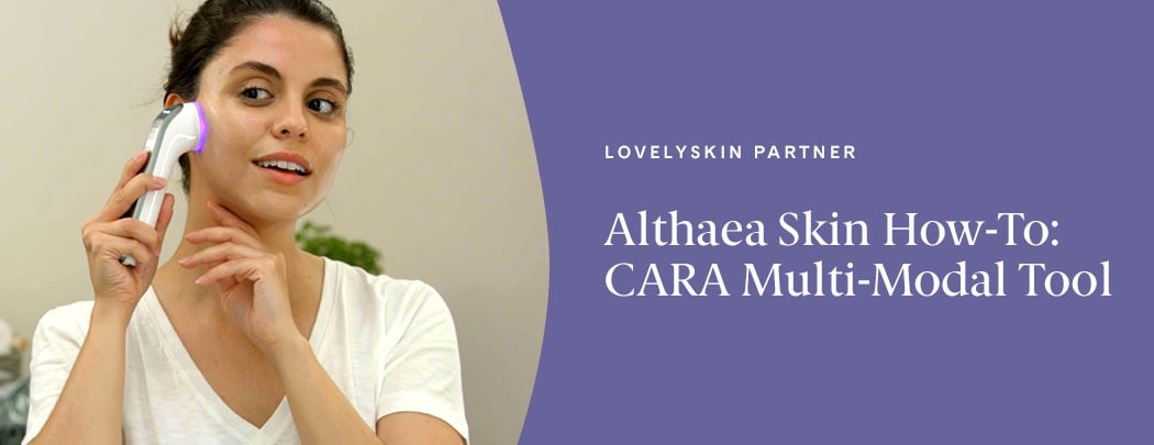 A woman with medium, olive-toned skin holds the Althaea Skin CARA Multi-Modal tool to her right cheek. She is in a bathroom setting.