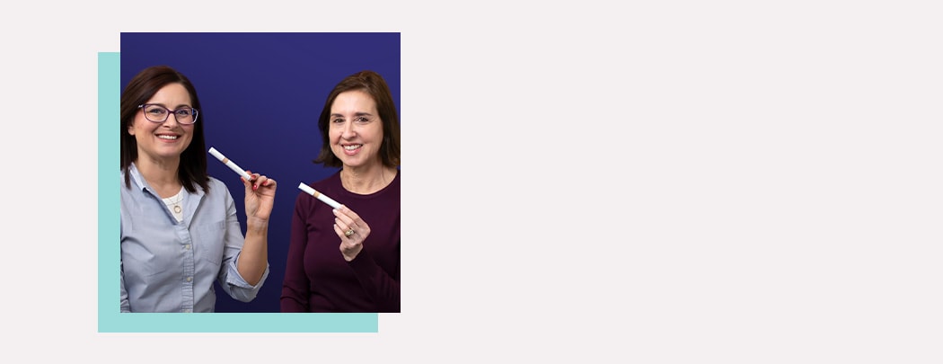 Two women, one in a dark purple shirt and one in a light blue shirt, hold Jane Iredale Skintuition Brightening Under Eye Concealer while smiling.