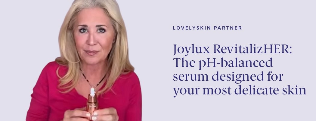 A woman with a medium skin tone, blonde hair and brown eyes looks at the camera while holding a bottle of Joylux revitalizHER Hydrating Serum.