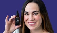 A woman with olive toned skin and dark brown hair smiles while holding a brown serum bottle that has a black label.