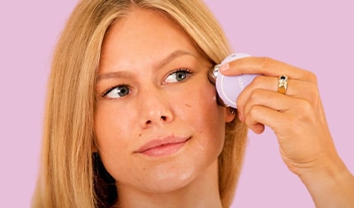 A woman with a light to medium skin tone and blonde hair presses a microcurrent device to her cheekbone.