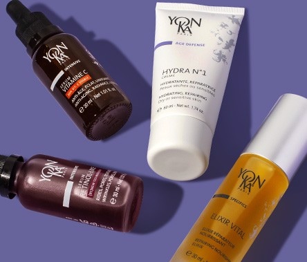 Four products, one in a white tube, two in clear glass bottles, and one in a brown glass bottle, lay on a medium purple background.