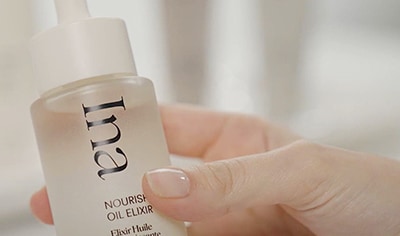 A person with fair skin and polished nails holds a bottle of Ina Nourishing Oil Elixir.