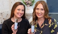 Two women with brown hair holding white bottles of skin care serum and smiling. A vase of flowers sits in between them.