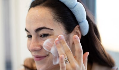A woman with a medium skin tone and dark brown hair wearing a light blue headband applies a foaming cleanser to her face with a soft smile.