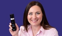A woman with fair skin and dark hair smiles while holding a dark bottle of SkinCeuticals Advanced RGN-6. She wears a pink shirt.
