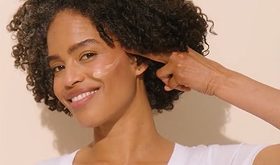 A woman with a medium to deep skin tone and brown curly hair presses a swatch of ISDIN Eryfotona Ageless Tinted Sunscreen SPF 50 to her cheek.