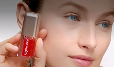 A person with fair skin holds a tube of Jane Iredale Just Kissed pH Lip Oil next to their cheek.