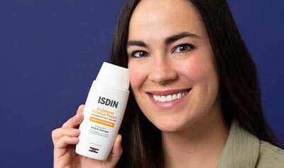 A woman with olive toned skin and dark hair holds a bottle of ISDIN Photo Eryfotona Actinica Tinted Daily Lightweight Mineral SPF 50+ while smiling.