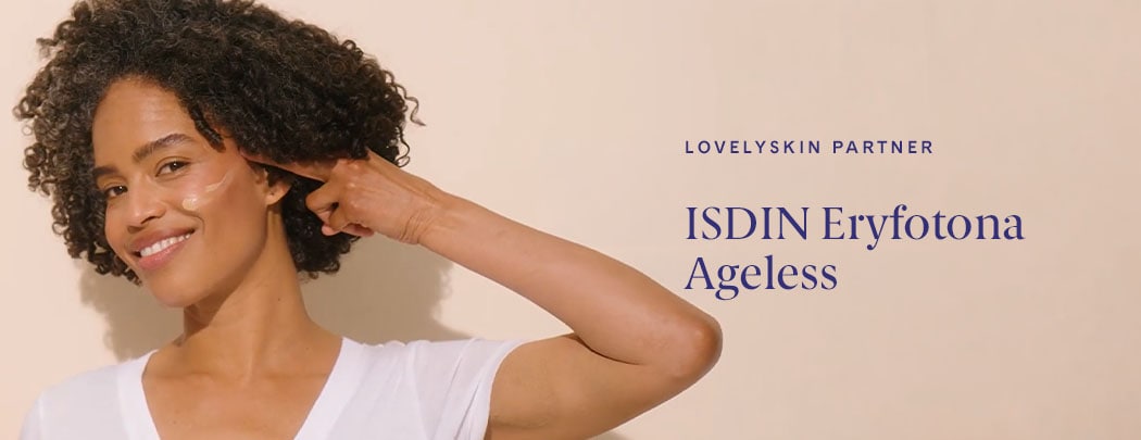 A woman with a medium to deep skin tone and brown curly hair presses a swatch of ISDIN Eryfotona Ageless Tinted Sunscreen SPF 50 to her cheek.