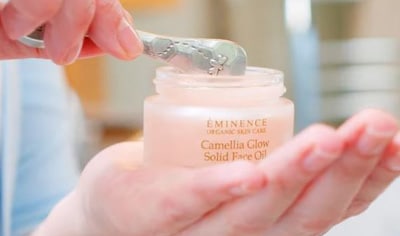 One hand holding a jar of Eminence Organics Camellia Glow Solid Face Oil while the other uses a metal spoon to scoop the product