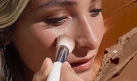 A woman with a light skin tone and blonde hair presses a brush to her cheek. There are swatches of the Ere Perez Cacao Bronzing Pot behind her.