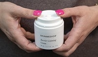A person with pink fingernails holds a jar of SkinMedica Barrier Calming Cream while pressing down on the airless pump jar to dispense product.