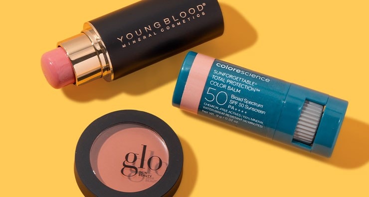 Blush sticks from Youngblood and Colorescience and a circular pot of blush by Glo all on an orange background.