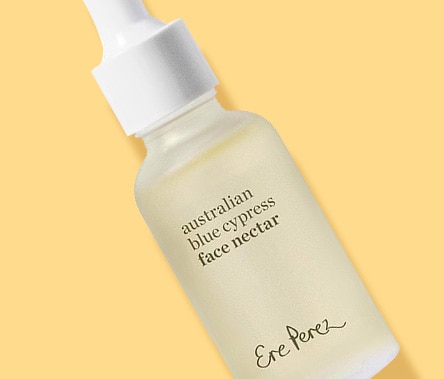A woman with a medium skin tone and short brown hair drops Ere Perez Australian Blue Cypress Face Nectar onto her cheek.