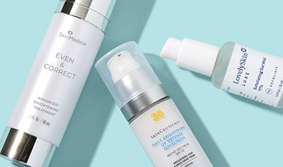 Bottles of SkinMedica Even & Correct serum, SkinCeuticals sunscreen and LovelySkin Exfoliating Gel on a light blue background.